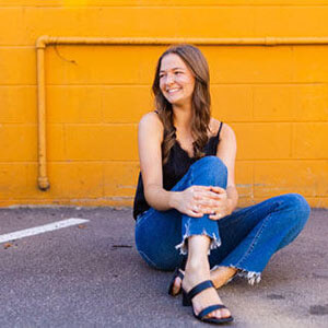 Kallie in a black top and blue jeans sitting against orange wall and wearing black sandals