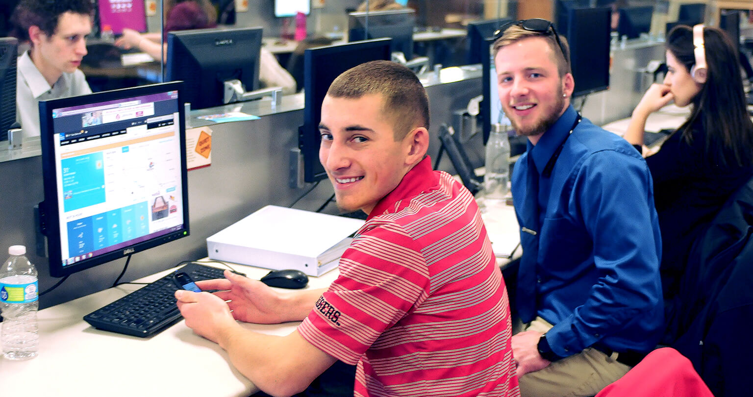 People working at computer stations in a classroom setting, one viewing a website interface on screen.
