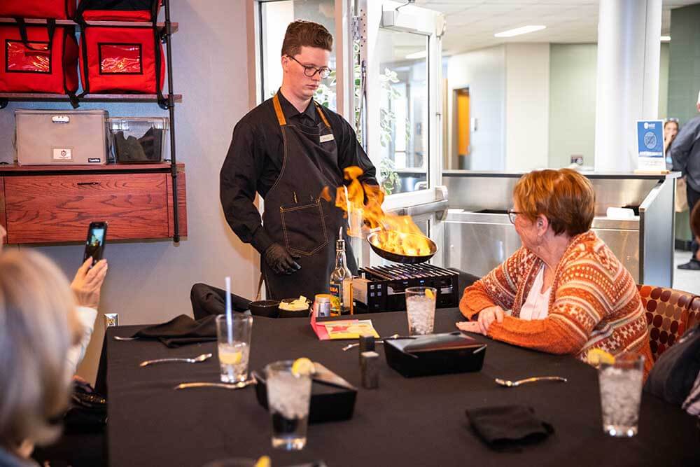 Cook prepares a flaming dish at a tabletop burner at 620 West Restaurant and Cafe while guests watch. Plates, drinks, and utensils are set on the table during the tableside cooking demonstration.