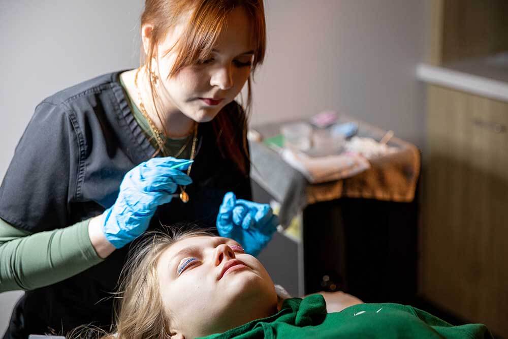 Aesthetician student wearing gloves performs a facial treatment on a client lying on a treatment bed in the 620 West Medspa and Massage Clinic. The practitioner works near the client's eye area using a small tool.