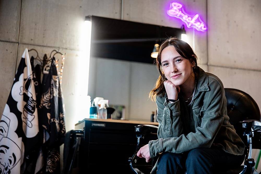 Brecken seated in a styling chair in a salon, leaning forward with chin resting on hand, facing the camera. A lit mirror, styling tools, patterned cloth, concrete wall, and purple neon sign are visible in the background.