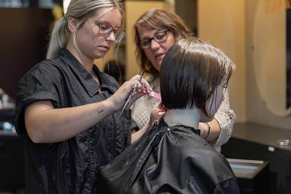 Cosmetology student cuts hair on a mannequin head while an instructor observes closely. Scissors, cape, and Shear Inspiration salon workspace are visible during hands-on training.