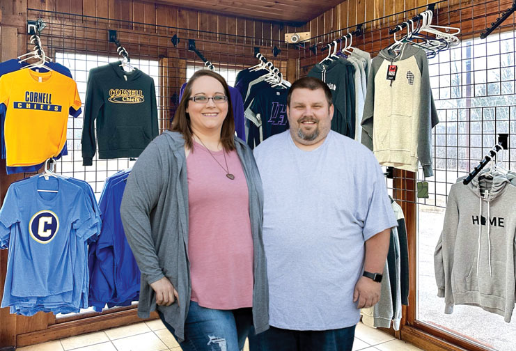 Jamie Boulding an Matt, her husband,e standing in a college apparel store, surrounded by hanging T-shirts, hoodies, and sweatshirts displayed on wall-mounted racks and window displays.