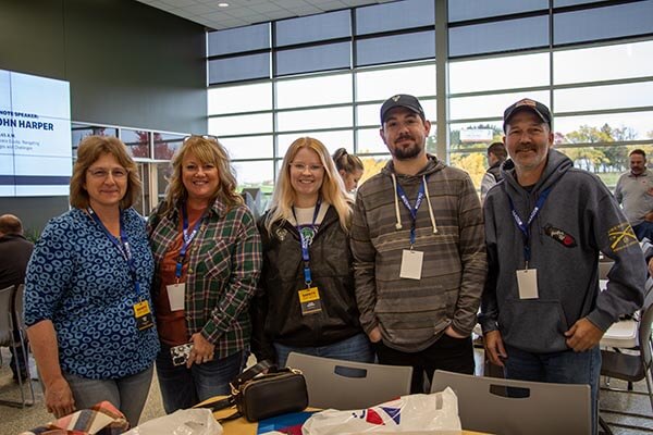 Five people with name tags pose indoors near tables and windows; bags and a 'John Harper' sign are visible.