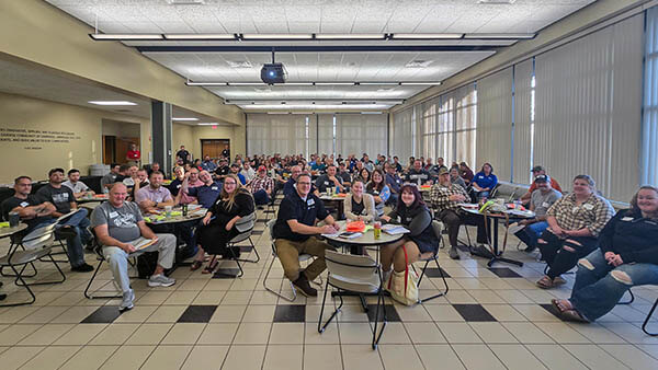 Large group seated at round tables in a bright conference room during a meeting or training session.