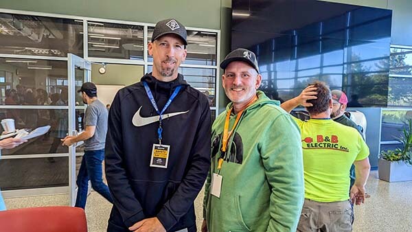 Two people with ID badges pose indoors; others in the background are talking, holding papers, or adjusting a hat.