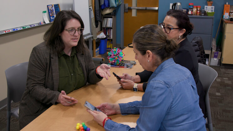 Three people discuss around a table with colorful objects, one holding a phone, another gesturing.