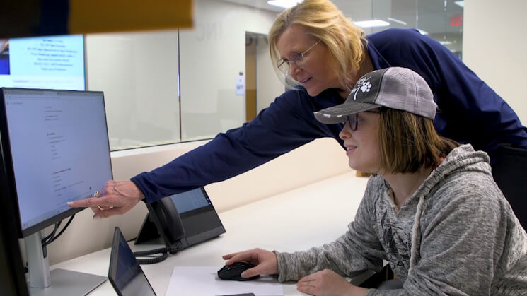 Person in blue shirt points at screen while another in gray hoodie and cap uses the mouse.