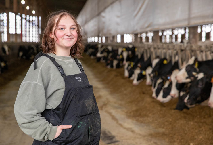Woman in overalls standing in a barn with cows behind her