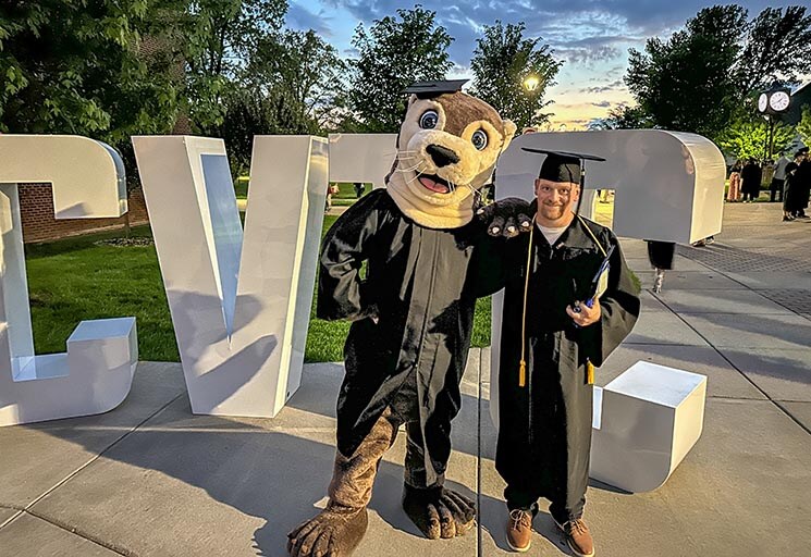 A graduate in a gown and cap poses with Ollie the Otter mascot in front of large CVTC letters. The sunset adds a warm glow to the outdoor scene.
