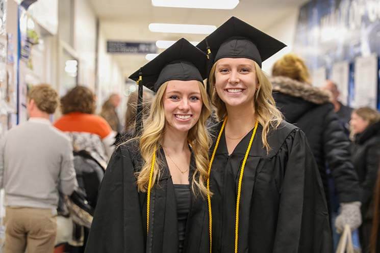 Two graduates in black gowns and caps with gold honor cords stand in a hallway. The background shows other people walking or standing.