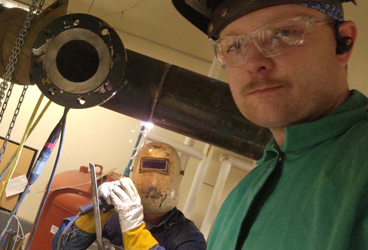 Two men, a father and son, weld together. One with a welding helmet on, and one with it tipped up to show his face.
