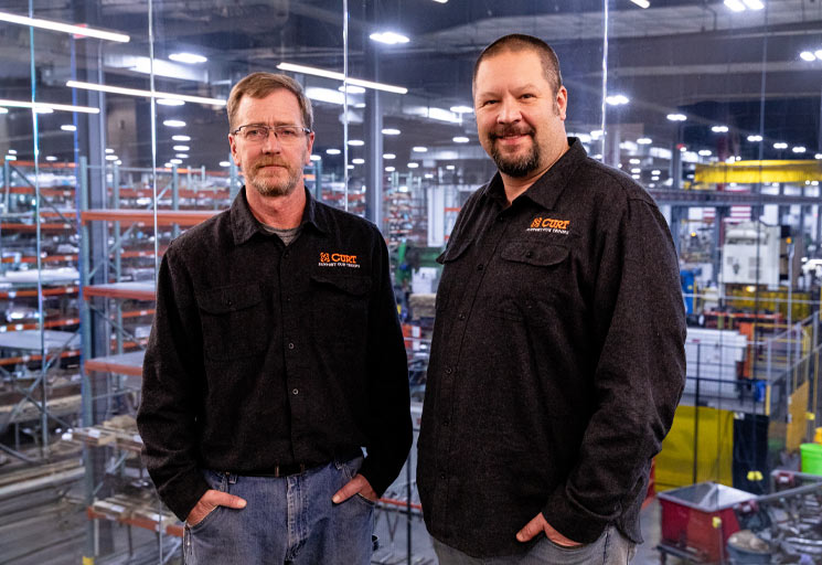 Two men posing in front of a window looking out over a manufacturing plant